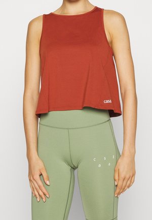 Vrouw draagt een mouwloos crop top in roestig rood en een hoog getailleerde saliegroene legging met kleine witte logo's op beide kledingstukken.