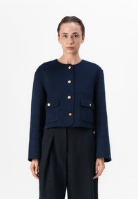 Jachetă din lână bleu marin cu guler rotund, mâneci lungi, închidere cu butoane aurii și două buzunare frontale. Design scurtat cu un finisaj texturat.
