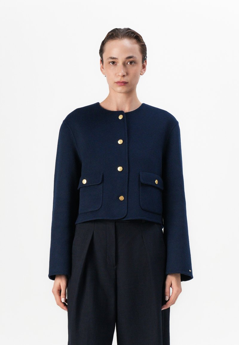 Jachetă din lână bleu marin cu guler rotund, mâneci lungi, închidere cu butoane aurii și două buzunare frontale. Design scurtat cu un finisaj texturat.