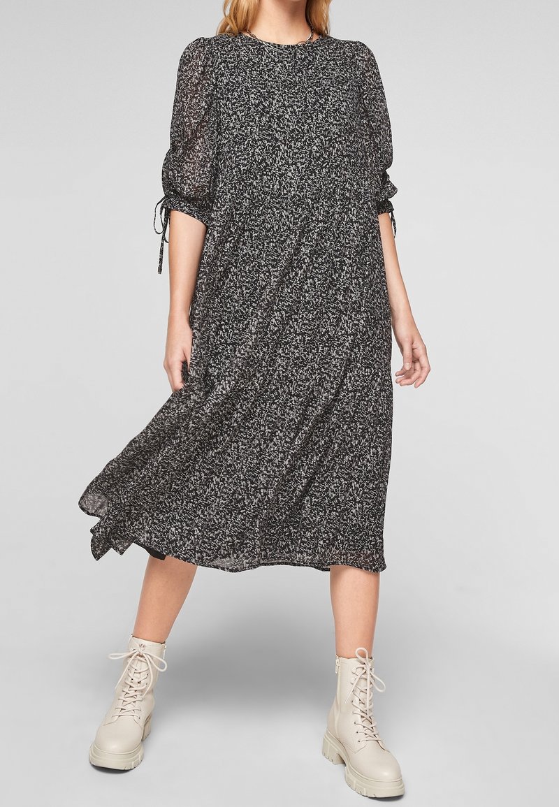 Femme portant une robe midi noire et blanche tachetée avec des manches 3/4 transparentes nouées aux coudes, associée à des bottines blanches à lacets.