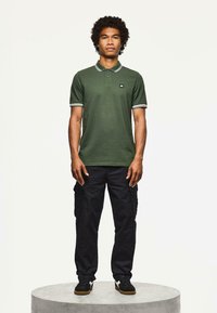 Giovane uomo in piedi su una piattaforma circolare grigia, con indosso una polo verde a maniche corte, pantaloni cargo neri e scarpe da ginnastica nere con strisce bianche.