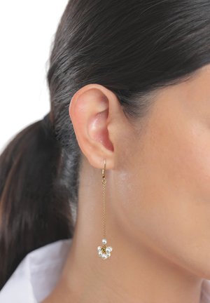 Gros plan sur l’oreille d’une femme aux cheveux foncés attachés, portant une longue boucle d’oreille chaîne en or avec un bouquet de petites perles blanches.