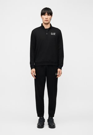 Ung person står iført sort lynlåstrøje og matchende sorte bukser med EA7-logo samt sorte sneakers på ensfarvet hvid baggrund.