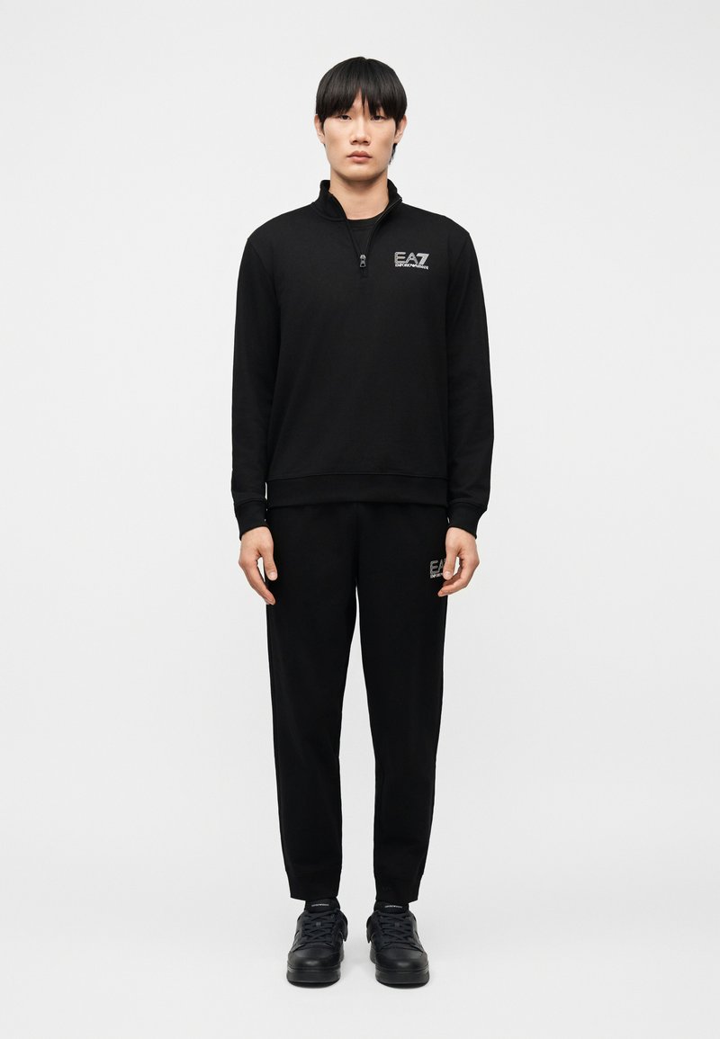 Νεαρό άτομο που στέκεται, φοράει μαύρο φούτερ με φερμουάρ και μαύρο παντελόνι με το λογότυπο EA7, καθώς και μαύρα αθλητικά παπούτσια, σε απλό λευκό φόντο.