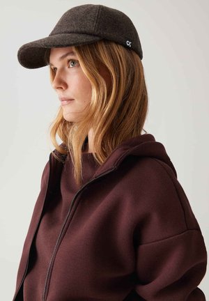 Braune Wollmütze mit einem gebogenen Schirm, kombiniert mit einer weinroten Zip-Hoodie. Die Textur wirkt weich und das Design ist lässig. Auffälliges Logo-Detail auf der Mütze.