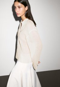 Blouse beige clair, semi-transparente, avec une coupe décontractée et des manches longues, associée à une jupe fluide de couleur crème. Texture lisse.