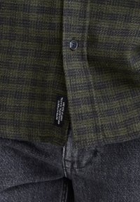 Nærbillede af en grøn og sort ternet skjorte, knappet over mørkegrå denimjeans med et sort mærke syet nær skjortens kant.