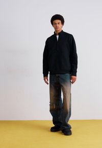 Sudadera negra con cremallera y cuello alto, combinada con jeans azules desgastados que presentan rodillas rasgadas, sobre un fondo blanco.