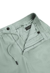 Pantalones verde claro con cintura elástica, cordón, cremallera y cierre con botón, mostrando la etiqueta de la marca en el interior de la cintura.