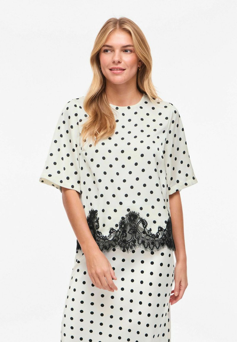 Blouse crème à pois noirs, manches courtes amples et ourlet décoratif en dentelle noire. Texture lisse avec un léger brillant.