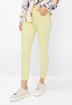 Person iført lysegule tætsiddende bukser, pastelmønstret bluse og lyserøde sneakers mod en ensfarvet hvid baggrund.