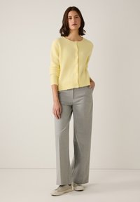 Gelber Wollcardigan mit rundem Ausschnitt und Knopfverschluss, kombiniert mit grauen, maßgeschneiderten Hosen und hellen Turnschuhen. Weiche Textur sichtbar.