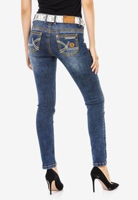 Schmale blaue Denim-Jeans mit Logo-Details und kontrastierenden Nähten. Ausgestattet mit einem silbernen Nieten-Gürtel. Tragen mit schwarzen Stilettos.