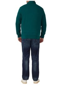 Groene sweatshirt met hoge kraag, gecombineerd met donkerblauwe jeans en witte schoenen. De sweatshirt heeft geribbelde manchetten en onderrand.