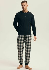 Mężczyzna stojący boso, ubrany w czarną koszulę henley z długim rękawem oraz czarno-białe kratkowane spodnie do piżamy na jednolitym tle.