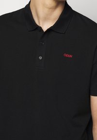 Muž v černém polo tričku s límečkem a třemi knoflíky, s červeným vyšitým textem na levé hrudi.