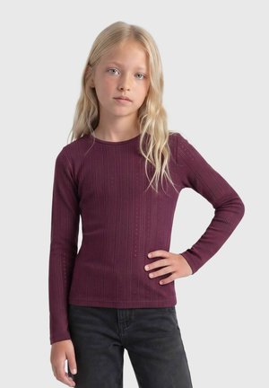 Fille blonde aux longs cheveux porte un haut violet à manches longues ajusté et un pantalon noir, posant avec une main sur la hanche.