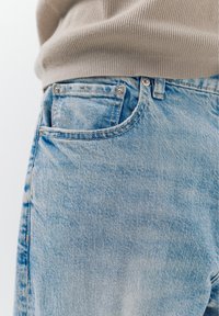 Svetlomodré riflové nohavice so viditeľným predným vreckom, striebornými nitmi a béžovým rebrovaným svetrom mierne zastrčeným v páse.