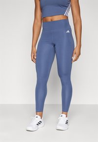 Blå träningsleggings med hög midja och slät textur. Har en vit Adidas-logotyp på höften och tre vita ränder längs sidorna.