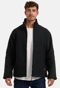 Chaqueta de forro polar negra con un alto collar, cierre de cremallera y bolsillos frontales. Presenta una textura suave y un branding sutil en el pecho.