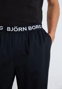 Calções pretos com um cós elástico, ostentando a marca "BJÖRN BORG" em branco. O tecido é suave, com um corte relaxado e bolsos laterais.