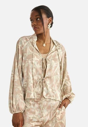 Mujer con chaqueta beige de estampado floral con lazos, pantalones a juego, collar con colgante dorado y pendientes de flores blancas, mirando hacia un lado.