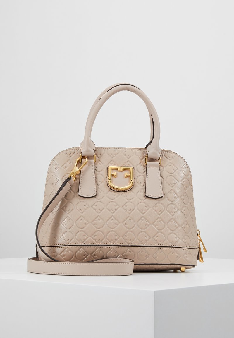 Sac à main en cuir beige avec motif géométrique embossé, plaque logo dorée, doubles poignées, bandoulière amovible et fermeture éclair dorée sur surface blanche.
