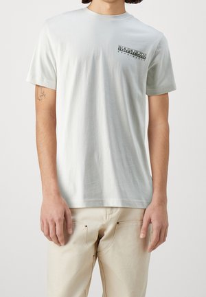 Man wearing light gray Napapijri Geographic t-shirt and off-white pants, standing with arms relaxed by sides against plain background.