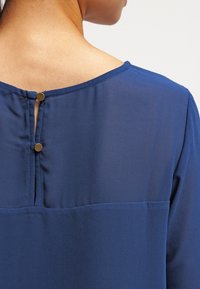 Mörkblå blus med en slät textur, rundad halsringning och två guldknappar bak för stängning.
