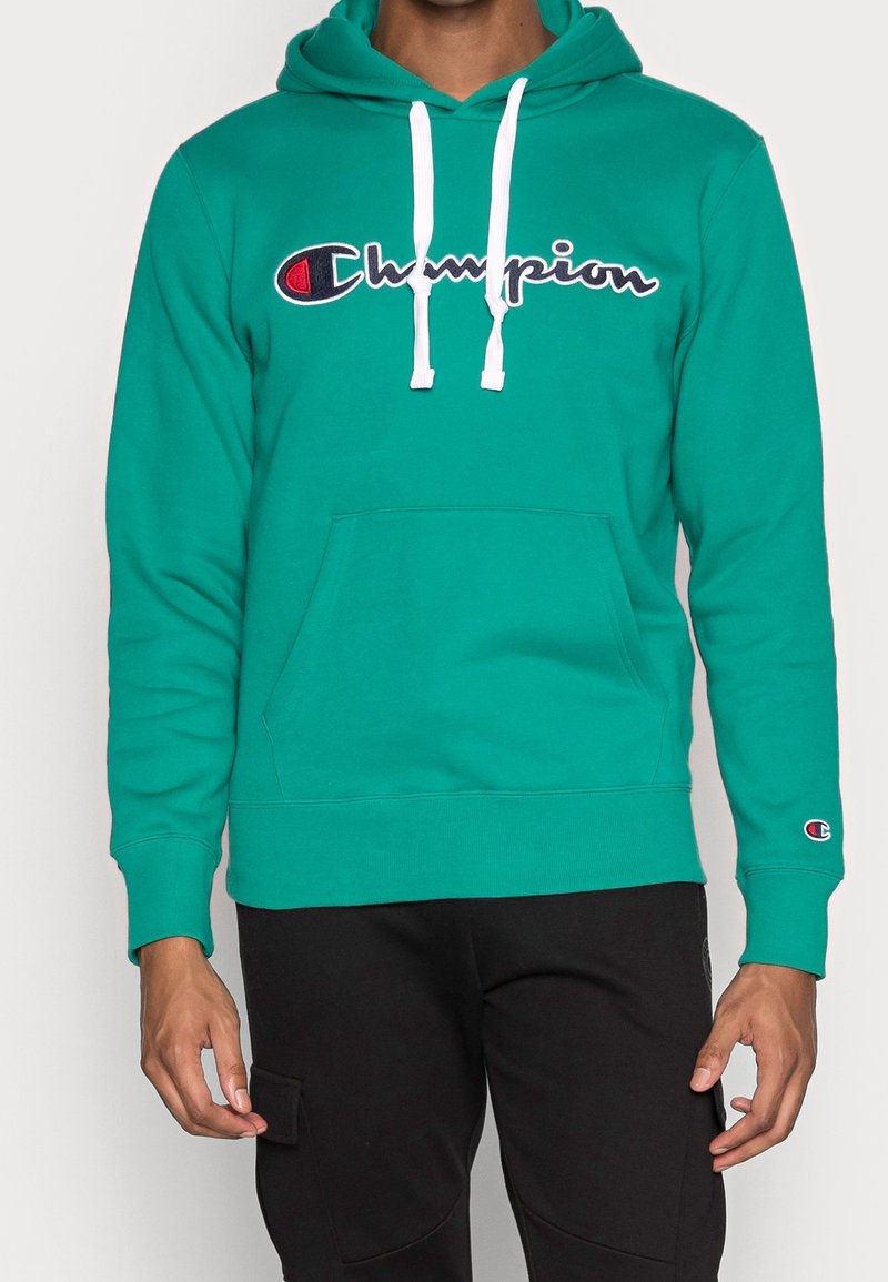 Vihreä huppari, valmistettu puuvillasekoitteesta, jossa on valkoiset kiristysnauhat ja etutasku. Champion-logo punaisena ja mustana rinnassa.