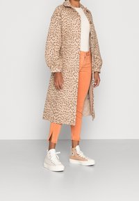 Manteau en imprimé léopard beige avec des manches ballons, associé à un pantalon orange à la cheville et des sneakers montants beiges, présentant des accents et des lacets blancs.