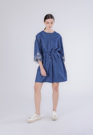Kobieta w niebieskiej sukience do kolan z haftowanymi szerokimi rękawami, zapięciem na guziki przy dekolcie, przewiązana w pasie, w białych tenisówkach, stojąca na jednolitym tle.
