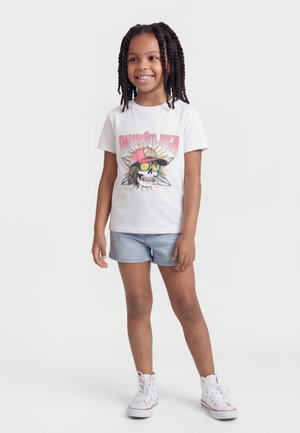 Naeratav tüdruk punutud juustega, kandes valget Quiksilveri T-särki värvilise kolju graafikaga, heledaid teksapükse ja valgeid tenniseid, seismas.