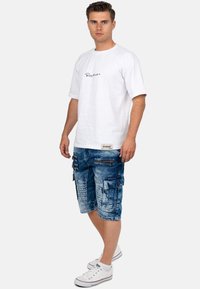 T-shirt bianco a maniche corte con logo nero, abbinato a pantaloni cargo in denim blu con molteplici tasche e cerniere, indossato con sneakers bianche.