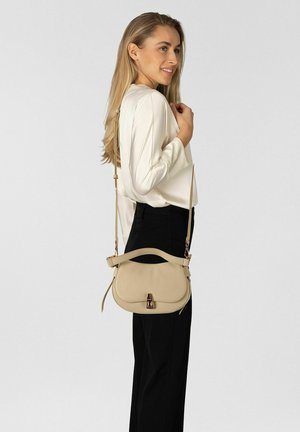 Mujer con cabello largo y rubio, vestida con una blusa blanca y pantalones negros, llevando un bolso de cuero beige con un cierre dorado.