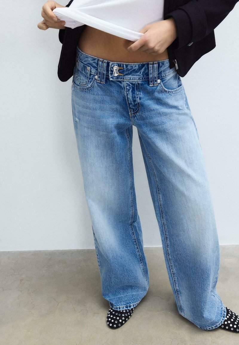 Person hebt weißes Hemd an, trägt hellblaue Jeans mit weitem Bein und schwarze Schuhe mit weißen Punkten, steht vor einer schlichten weißen Wand.