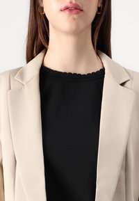 Béžové sako s vykrojenými klopami nošené přes černý top se skládaným výstřihem. Jsou vidět zlaté náušnice kruhy.