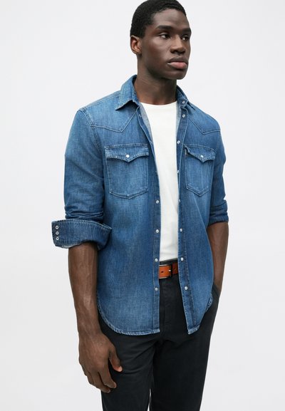 Camicia di jeans blu con due tasche sul petto, chiusura con bottoni e maniche arrotolabili. Abbinata a una maglietta bianca e pantaloni neri.