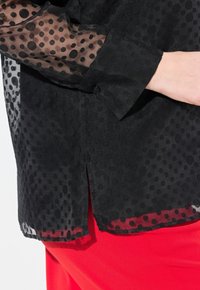 Blusa nera con maniche a pois trasparenti, dotata di una chiusura a bottoni e di un orlo spaccato. Indossata sopra un tessuto rosso brillante.