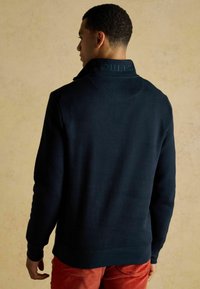 Laikahousuilla oleva tummansininen huppari, jossa on korkea kaulus ja kohokuviolla oleva brändinimi takana, valmistettu pehmeästä kankaasta, yhdistettynä punaisiin samettihousuihin.