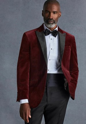 Burgundska baršunasta tuxedo jakna s crnim satenastim reverima, nosi se preko bijele plisirane košulje i crne leptir-mašne, u kombinaciji s crnim hlačama.