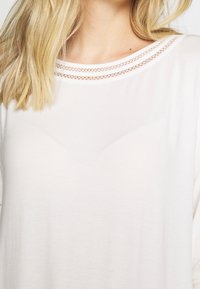 Blonde Frau, die ein weißes Oberteil mit doppelter Spitzenborte am Ausschnitt trägt und ihren Schlüsselbeinen und oberen Brustbereich zeigt.