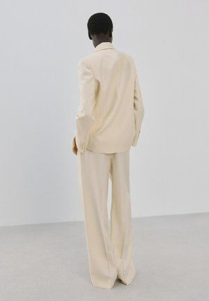 Osoba stojąca tyłem, ubrana w kremowy garnitur z szerokimi spodniami na tle jednolitego jasnoszarego tła.