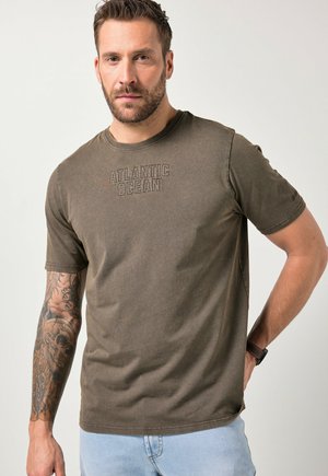 Uomo con capelli corti e barba, che indossa una t-shirt marrone con la scritta "Atlantic Ocean" e jeans azzurri, con un tatuaggio su tutto il braccio e un orologio da polso.