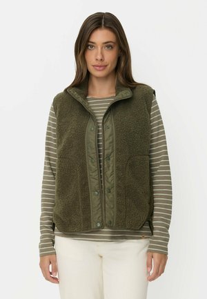 Woman wearing an olive green fleece vest over a striped long-sleeve shirt and white pants, standing against a plain background.
