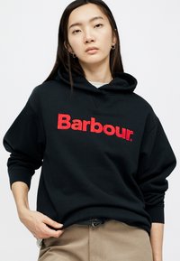 Noor inimene, kellel on pikad tumedad juuksed, kannab musta kapuutsi punase "Barbour" logoga, beeže pükse ja rõngakujulisi kõrvarõngaid, seisab valgel taustal.