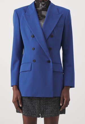 Kvinde iført en blå dobbeltradet blazer med sorte knapper over en sort mønstret top og et gråt tweedskørt med lynlås foran.