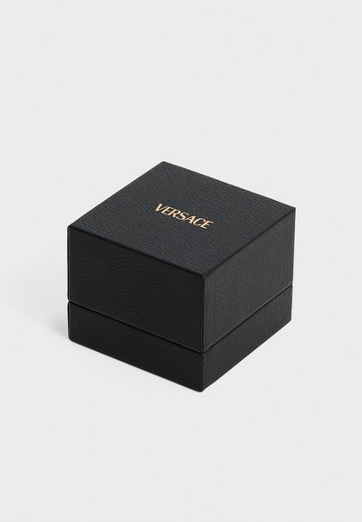 Boîte carrée texturée noire avec logo Versace doré centré sur le couvercle, posée sur un fond clair uni.