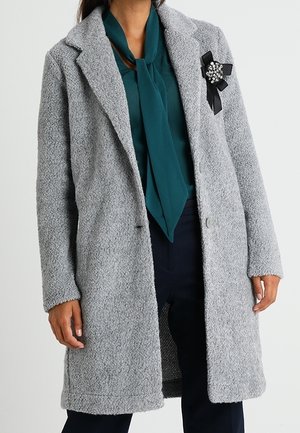 Femme portant un manteau gris clair texturé sur un chemisier bleu sarcelle avec un col à nouer et un pantalon foncé, avec une broche décorative en ruban noir sur le manteau.