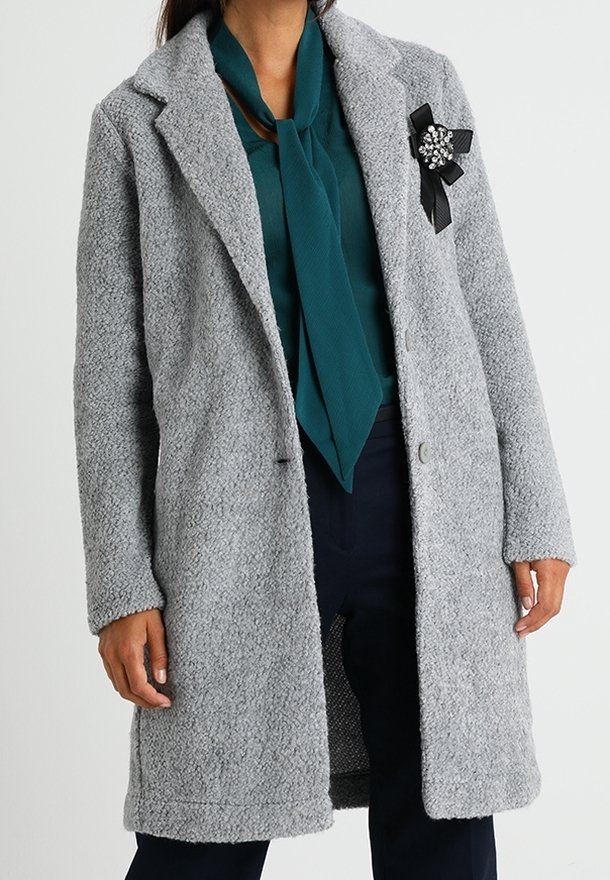 Femme portant un manteau gris clair texturé sur un chemisier bleu sarcelle avec un col à nouer et un pantalon foncé, avec une broche décorative en ruban noir sur le manteau.